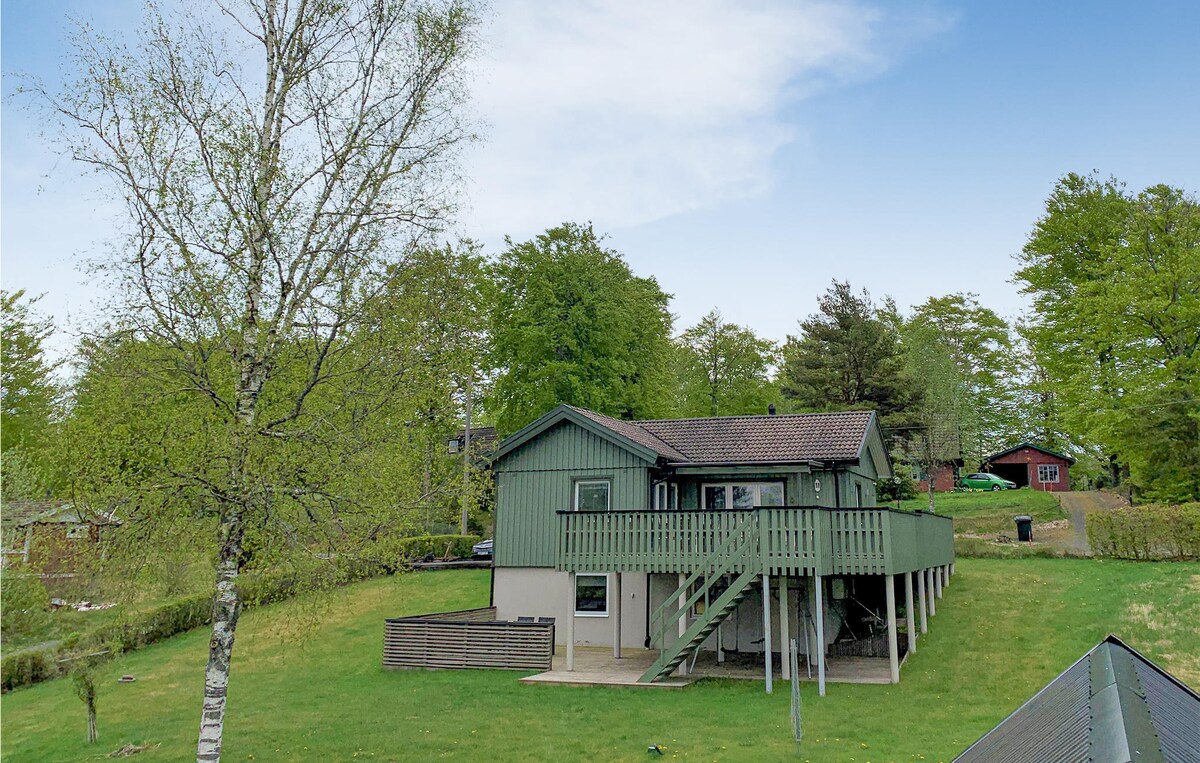 A charming house is set against a backdrop of greenery, featuring a spacious wooden terrace that extends from the upper level. The structure is surrounded by a well-kept lawn, providing ample outdoor space. Nearby trees enhance the serene environment.
