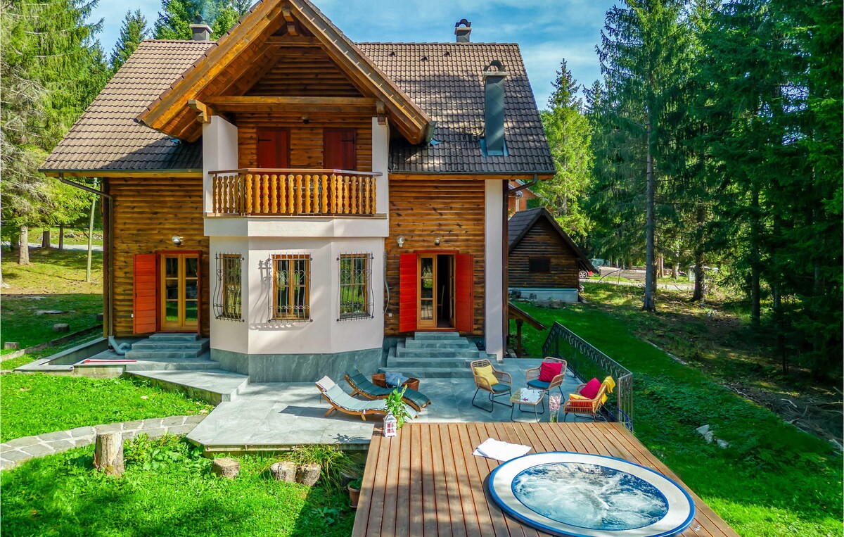 A charming two-story wooden holiday home is surrounded by lush greenery. The entrance features a spacious patio with access to a whirlpool, lounge chairs, and a balcony above, providing ample outdoor space for relaxation.