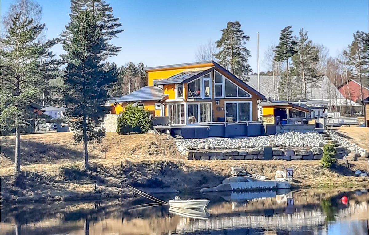 The house features a modern design with a combination of large windows and bright orange accents, set against a serene waterfront. A small boat is visible on the calm water, and the surrounding landscape includes sparse trees and well-maintained grounds.