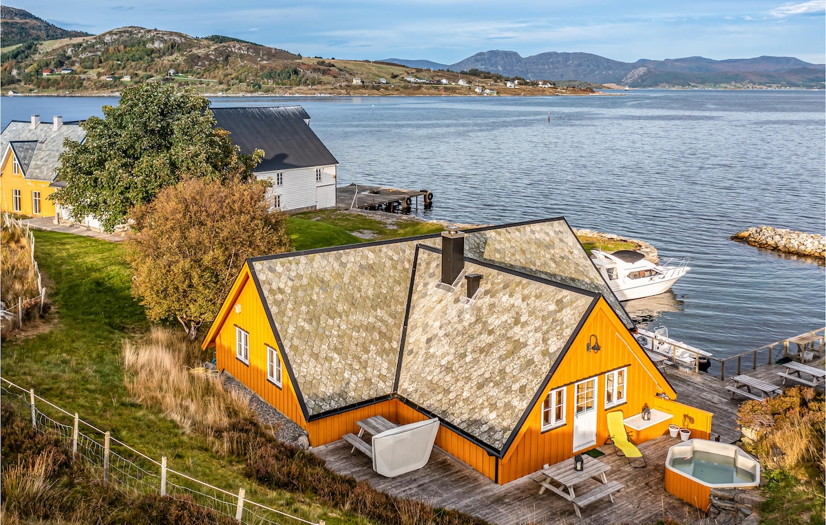 A vibrant orange house is situated by the water's edge, featuring a sloped roof and a spacious deck. Surrounding greenery and a neighboring building can be seen in the distance, along with boats docked nearby. The tranquil bay reflects the surrounding landscape.