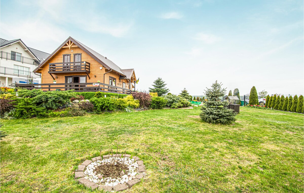 The exterior of a spacious vacation home is set within a well-maintained garden featuring lush green grass and neatly trimmed shrubs. A fire pit with a circular stone border is visible in the foreground, and the house is framed by trees, enhancing the tranquil rural atmosphere.