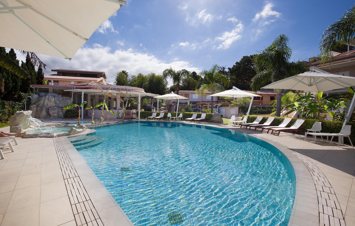 A large outdoor pool is surrounded by sun loungers and umbrellas. Lush greenery and palm trees can be seen in the background, complemented by a clear blue sky. A jacuzzi is positioned to one side, creating a serene atmosphere for relaxation.