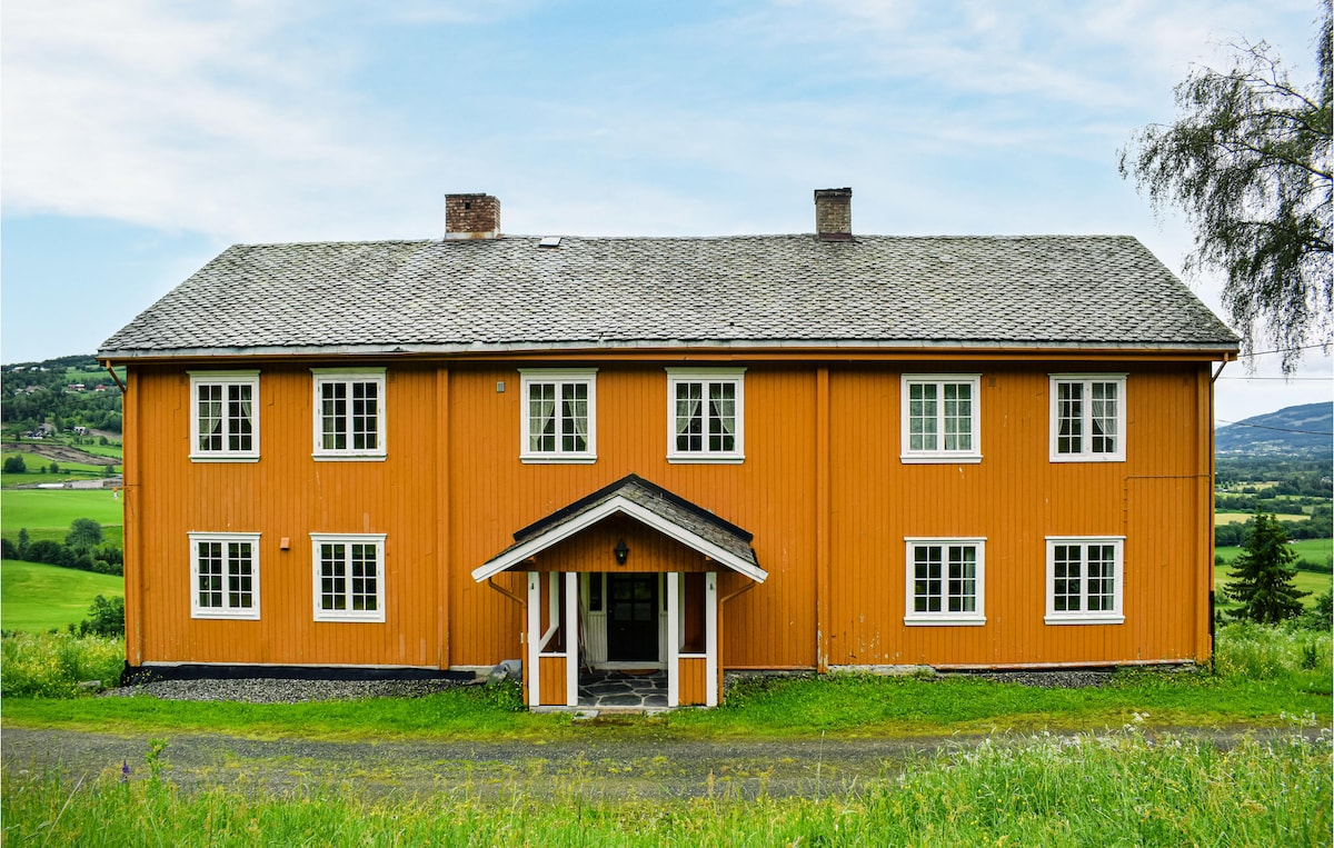 The farmhouse features a vibrant orange exterior, complemented by large white-framed windows. A welcoming entrance with a covered porch leads to the interior, while the surrounding landscape displays green fields and distant hills under a clear sky.