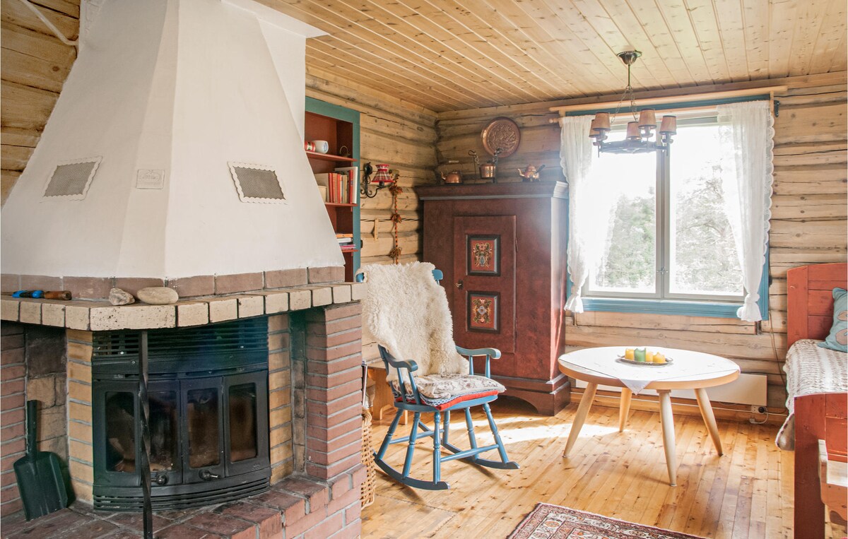 A cozy living space features a stone fireplace with a wood-burning stove, surrounded by logs. A comfortable rocking chair and a coffee table sit on a wooden floor. Natural light enters through a large window, flanked by sheer curtains, enhancing the tranquil ambiance.