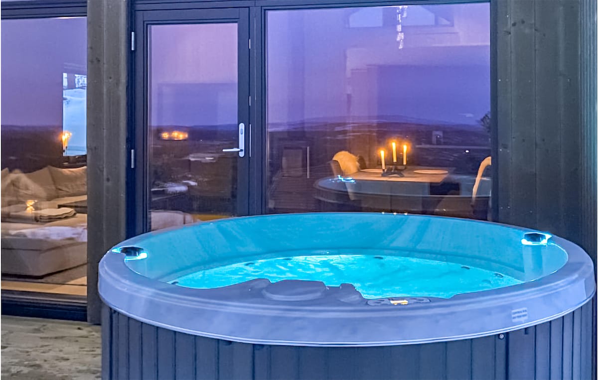 A round hot tub is positioned on the outdoor terrace, featuring glowing blue water. Large glass doors provide a view into the cozy interior, where soft lighting from candles creates a warm ambiance. The surrounding mountain landscape is visible in the twilight.