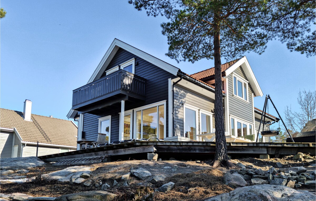A wooden house in classic Scandinavian style is elevated on rocky terrain, featuring large glass windows and a balcony overlooking the water. The exterior is painted in a deep blue, with a white trim. Lush trees surround the property, enhancing its serene setting.