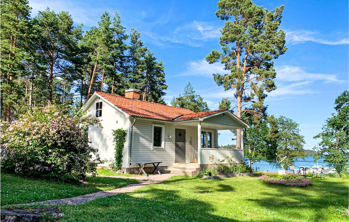 A charming vacation home is set amidst lush greenery, showcasing a light-colored exterior and a red-tiled roof. Surrounding the house are tall trees and blooming bushes, while a path leads to a spacious yard with outdoor seating, all with a view of the nearby water.