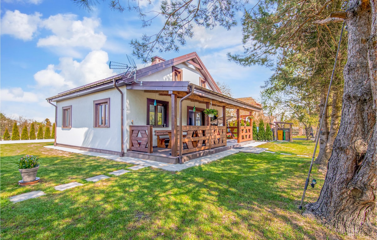 A charming vacation home is displayed, featuring a covered terrace with wooden columns and seating. The property is surrounded by well-maintained green grass and trees, providing a peaceful backdrop. The exterior showcases simple lines and light-colored walls, blending harmoniously with nature.