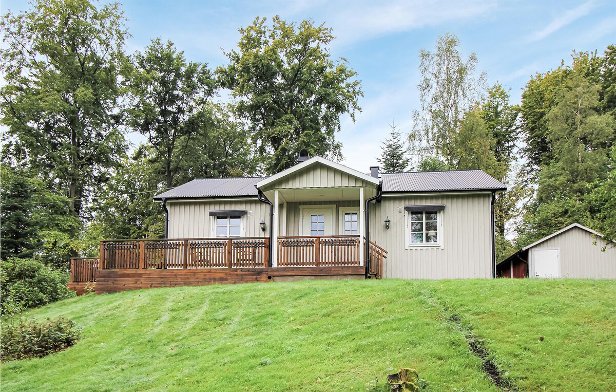 A charming cottage is nestled on a green hillside, surrounded by trees. The large wooden deck extends across the front, providing ample space for outdoor seating. The light-colored exterior is complemented by a dark roof, and a small outbuilding is visible to the right.