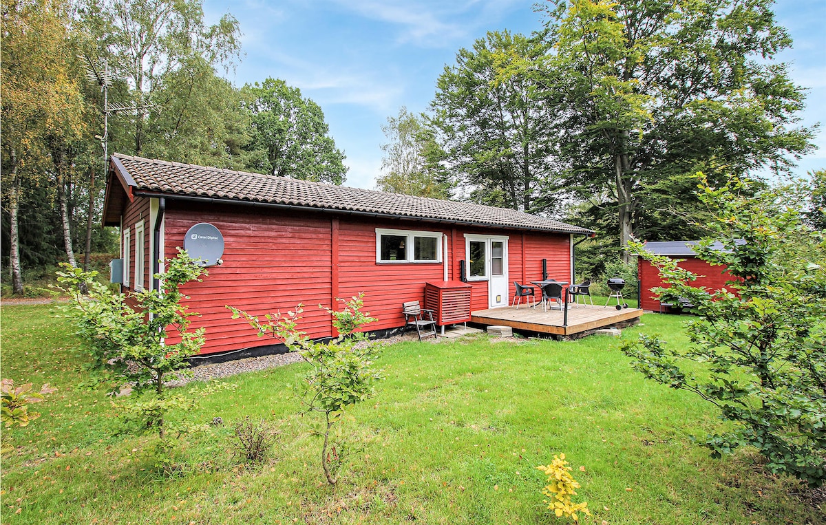 A cozy red house situated in a lush green environment, featuring a wooden deck with outdoor seating. The surrounding area includes well-maintained grass and trees, offering a natural setting that invites relaxation and outdoor activities.