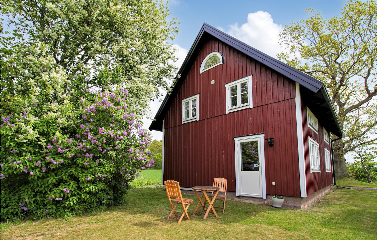 A charming red cottage is surrounded by lush greenery and flowering plants. Two wooden chairs and a small table are positioned on the grass in front of the entrance. Large windows reflect the natural light, enhancing the inviting exterior.