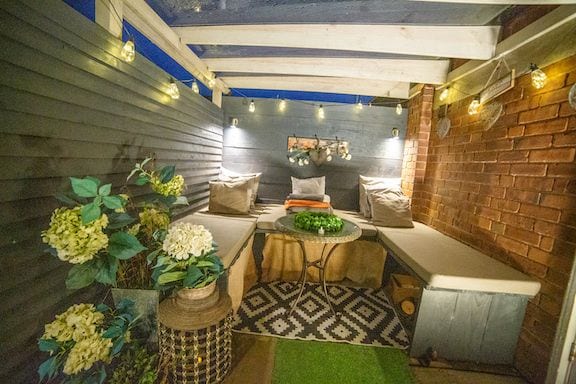 A cozy outdoor seating area is enclosed by wooden walls, illuminated by string lights. Comfortable cushions are placed on the benches, while a round table holds decorative items. Fresh flowers and greenery enhance the inviting atmosphere, completed by a patterned rug underfoot.