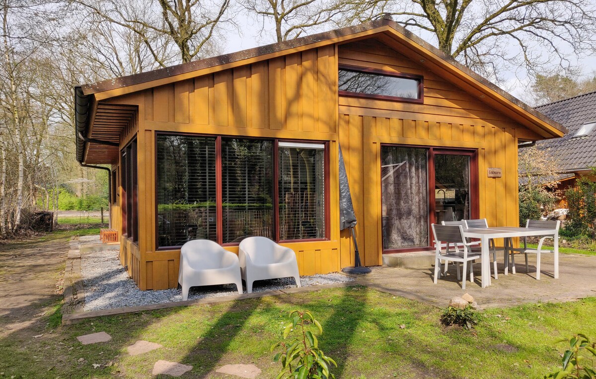 A wooden cottage showcases large windows, allowing natural light to fill the space. Two comfortable chairs are positioned outside, along with a table, highlighting an inviting area for relaxation. A well-kept garden surrounds the cottage, providing a serene atmosphere.