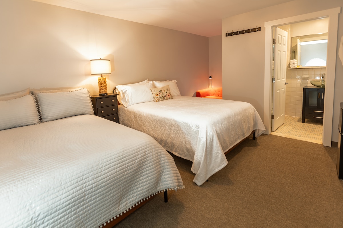 The room features two full beds dressed in white linens, complemented by a bedside table and lamp. A transitional space leads to an en suite bathroom, visible through an open door with modern fixtures and a neutral color palette.