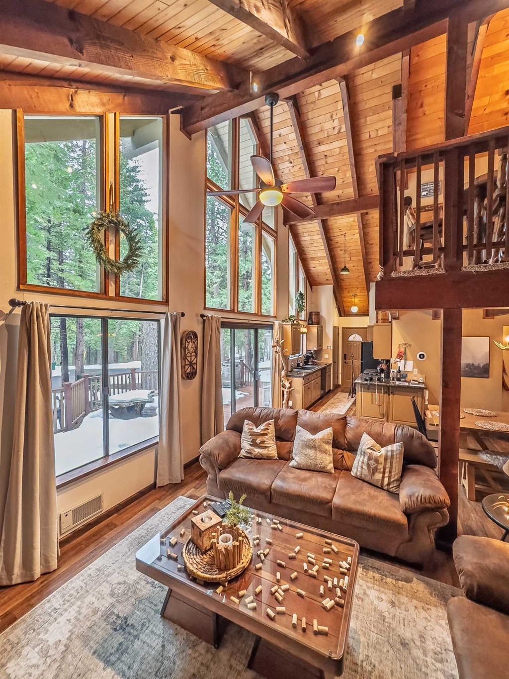 A spacious living room features high, vaulted ceilings with wooden beams and large windows that invite natural light. Comfortable leather sofas surround a wooden coffee table, which is adorned with decorative items. A view of the balcony is visible through the glass doors, enhancing the cozy ambiance.
