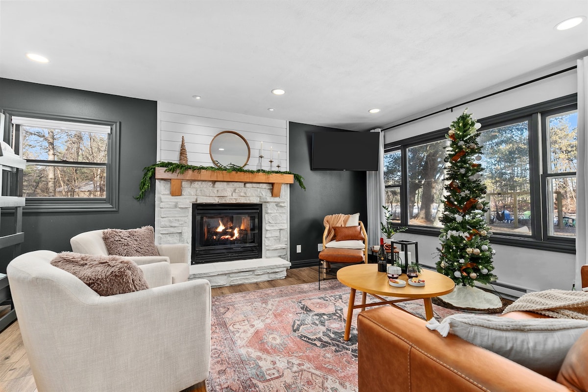 The inviting living area features a wood-burning fireplace framed by a stone surround, providing a cozy focal point. Large windows bathe the space in natural light, while a festive Christmas tree adds a seasonal touch. Comfortable seating invites relaxation in this warm setting.