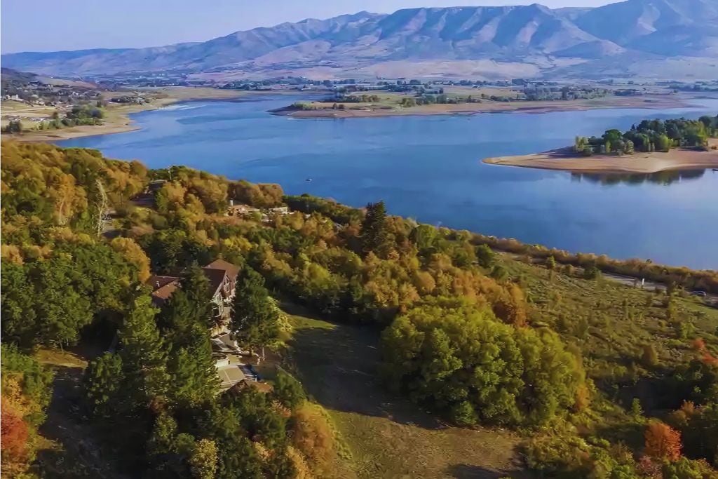Aerial view captures the serene landscape surrounding Lakeview Haven, showcasing lush trees and the expansive Pineview Reservoir. The mountains frame the horizon, and the vibrant colors of nature create a tranquil and inviting outdoor environment.