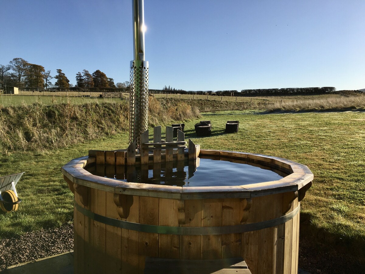 A wooden hot tub is situated in a grassy outdoor area, with sunlight reflecting off the warm water. Rolling fields and sparse trees create a serene backdrop, while a few lounge chairs are positioned nearby, inviting relaxation in the tranquil setting.