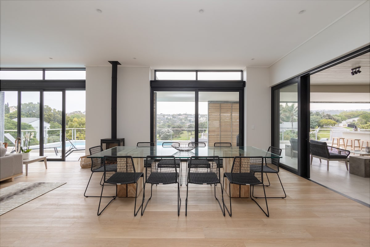 A modern dining area features a large glass table surrounded by eight black wire chairs. The space is illuminated by natural light from floor-to-ceiling windows, offering views of the outdoor area and surrounding landscape. A cozy seating arrangement is located nearby.