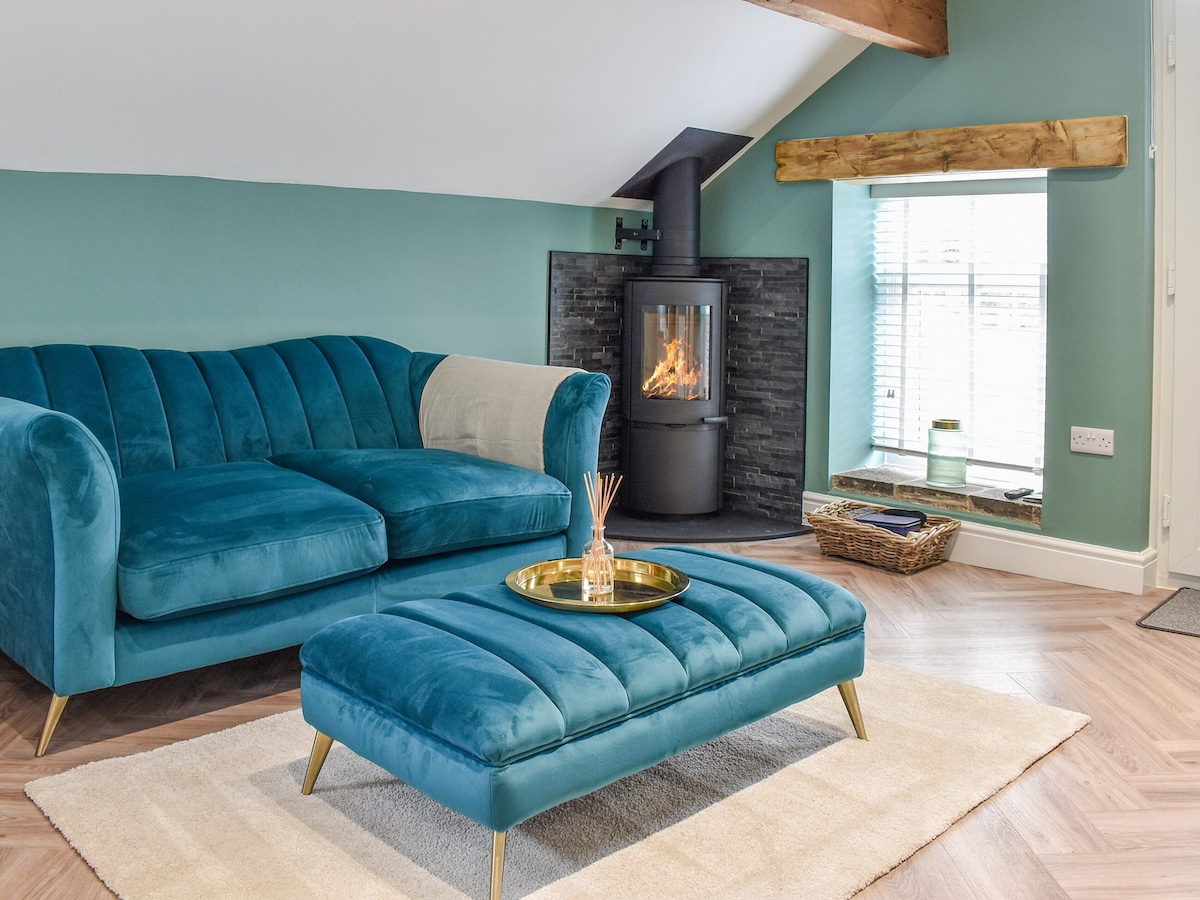 A comfortable living area is highlighted by a plush teal sofa and an ottoman with a metallic tray. A woodburner is positioned against a textured black wall, with flames visible. Natural light filters through windows covered by wooden blinds, creating a cozy ambiance.