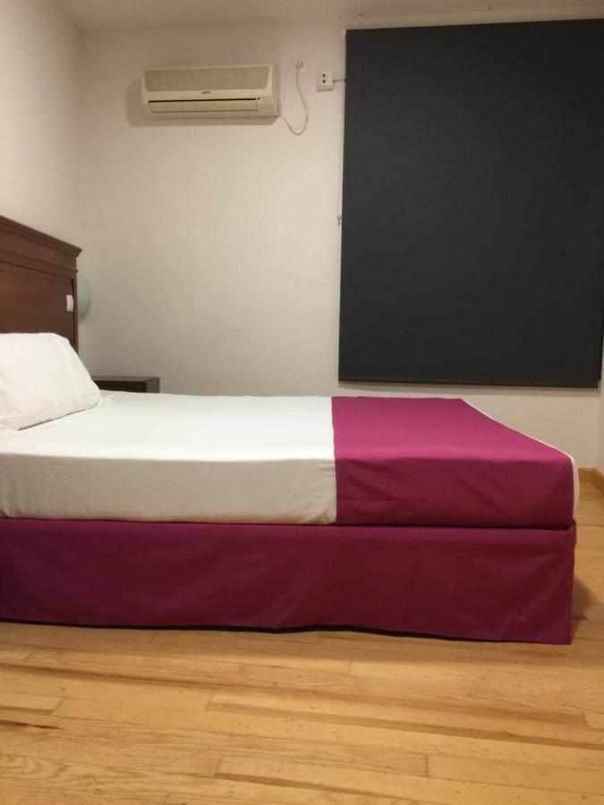 A cozy room features a double bed with a two-toned bedspread, a wooden nightstand with a lamp, and a wall-mounted air conditioner above. A large window is covered by a dark curtain, allowing for privacy and light control.