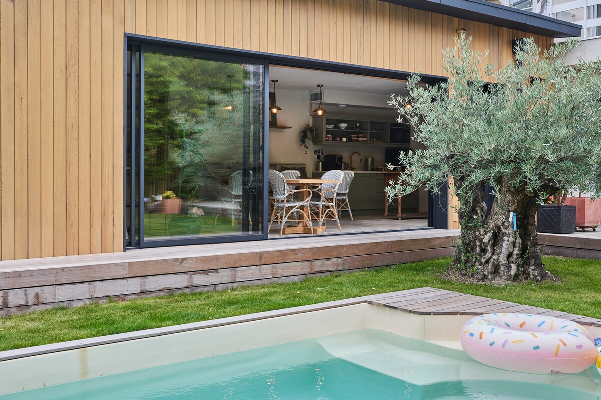 A modern building constructed with wooden panels is visible, featuring large glass doors that create a seamless connection to the outdoor space. A swimming pool reflects the surrounding greenery, while an olive tree adds to the tranquil atmosphere.