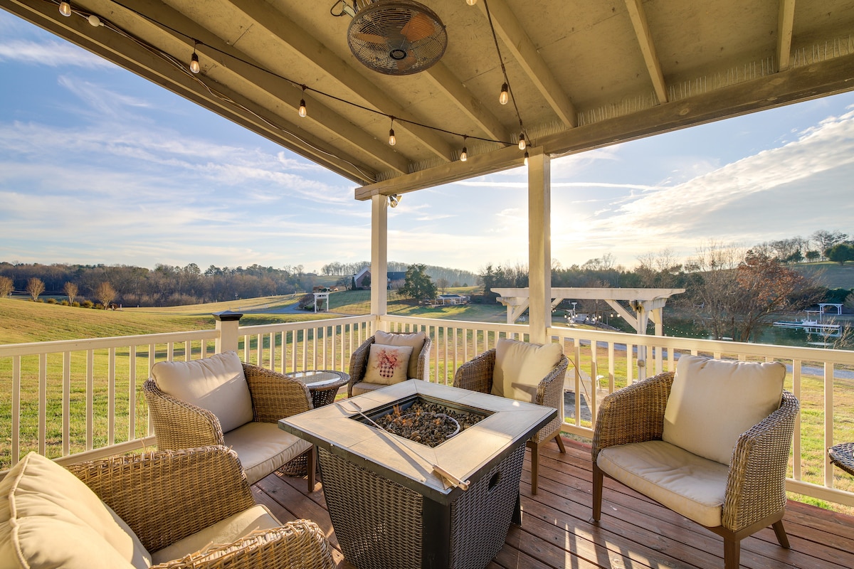 A cozy outdoor seating area is arranged around a central fire pit, featuring comfortable wicker chairs with soft cushions. Soft string lights are hung overhead, illuminating the space. A scenic view of rolling hills and open fields is visible in the background.