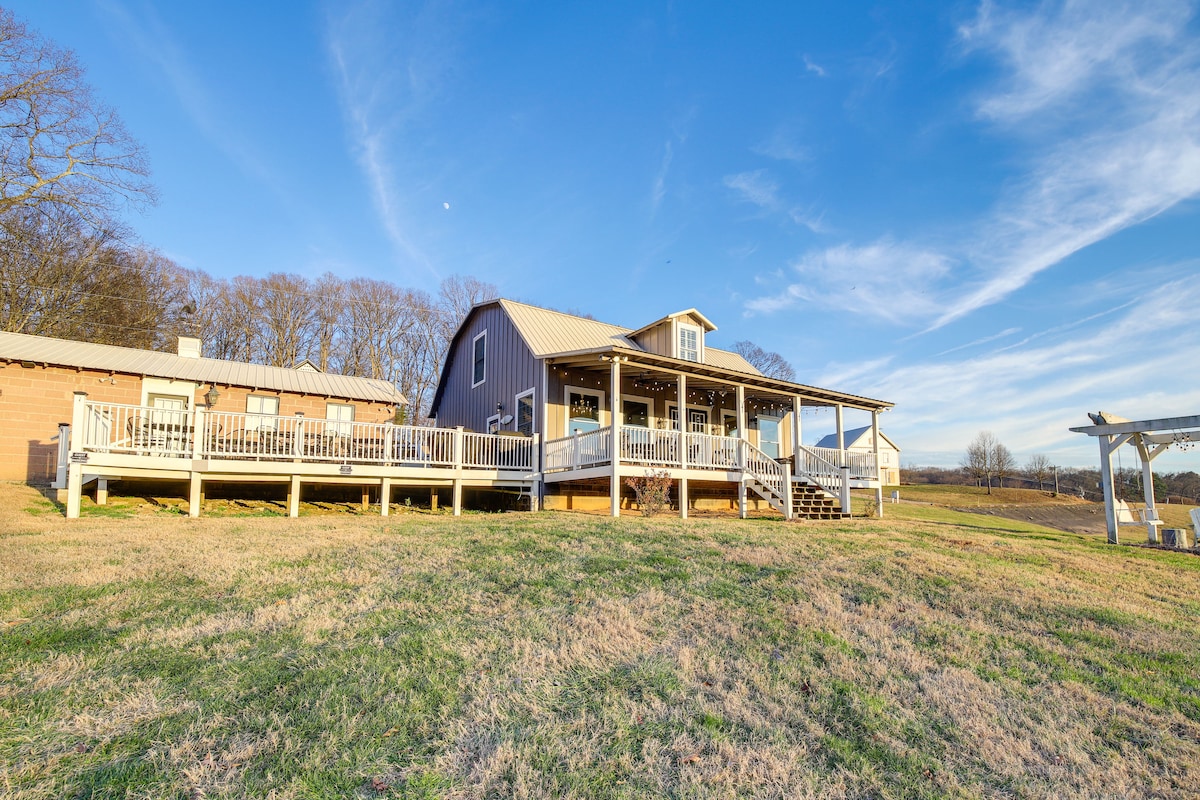 A spacious modern barn is positioned on a gently sloping grassy hill, featuring a wide deck that offers outdoor seating. The clear blue sky and soft clouds enhance the serene atmosphere, while trees line the background, providing a natural border to the property.