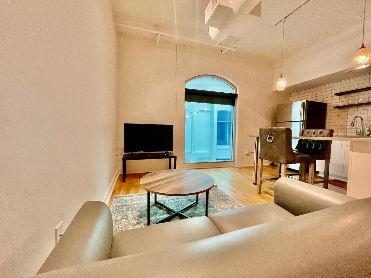 The living area features a comfortable beige sofa facing a wall-mounted television. A round wooden coffee table sits on a patterned rug, and a bar area with high stools is visible in the background. Natural light enters through an arched window overlooking the atrium.