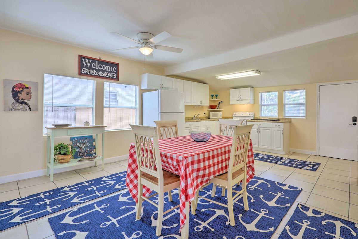 A bright dining area features a checkered red and white tablecloth covering a wooden table surrounded by four chairs. The space is furnished with white cabinets and appliances, while an anchor-patterned rug adds a coastal touch to the tiled floor. A welcome sign adorns the wall.