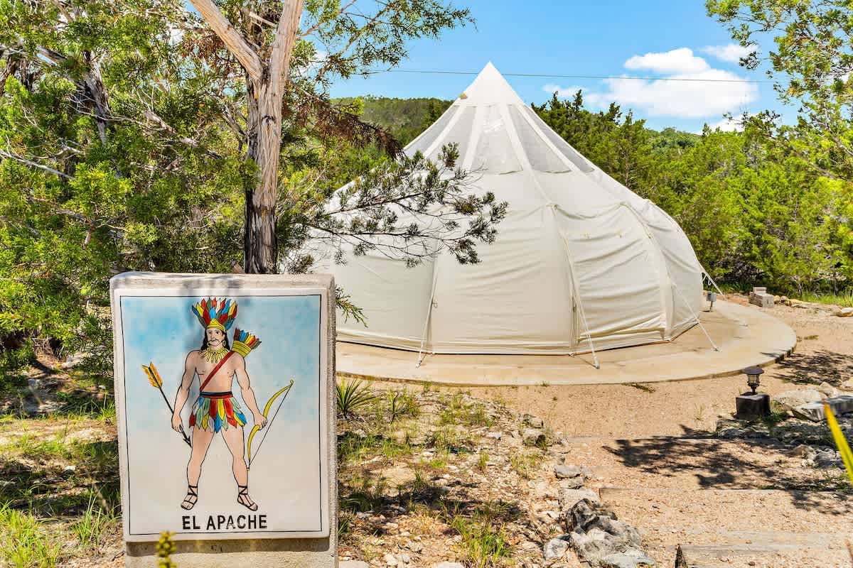A white glamping tent, featuring a pointed top, is surrounded by natural greenery. A colorful sign depicting a figure with arrows is positioned nearby, indicating the tent's name, 'El Apache.' A gravel path leads to the entrance, enhancing the rustic setting.