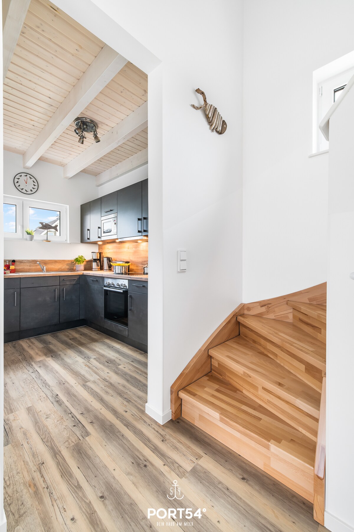 The image depicts a modern kitchen featuring dark cabinetry and wooden accents. Natural light is captured through windows, highlighting the clean lines of the space. A staircase with polished wooden steps leads to the upper level, enhancing the openness of the layout.