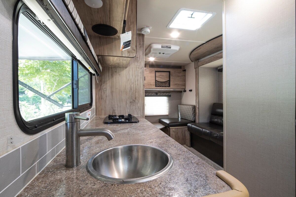 A well-equipped kitchen area is featured, with a stainless steel sink and a gas stove. Light-colored cabinetry complements the countertop. Large windows provide natural light and views of the surrounding greenery. A cozy seating area with a dark sofa is visible in the background.