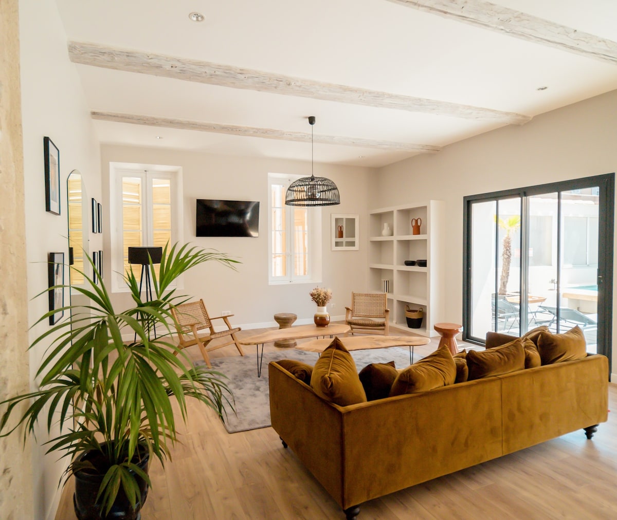 A spacious living room is highlighted by a large brown sofa and a central coffee table. Light wood beams add character to the ceiling, while a flat-screen TV and shelving unit complete the space. Large windows provide views of the outdoor area, enhancing natural light.