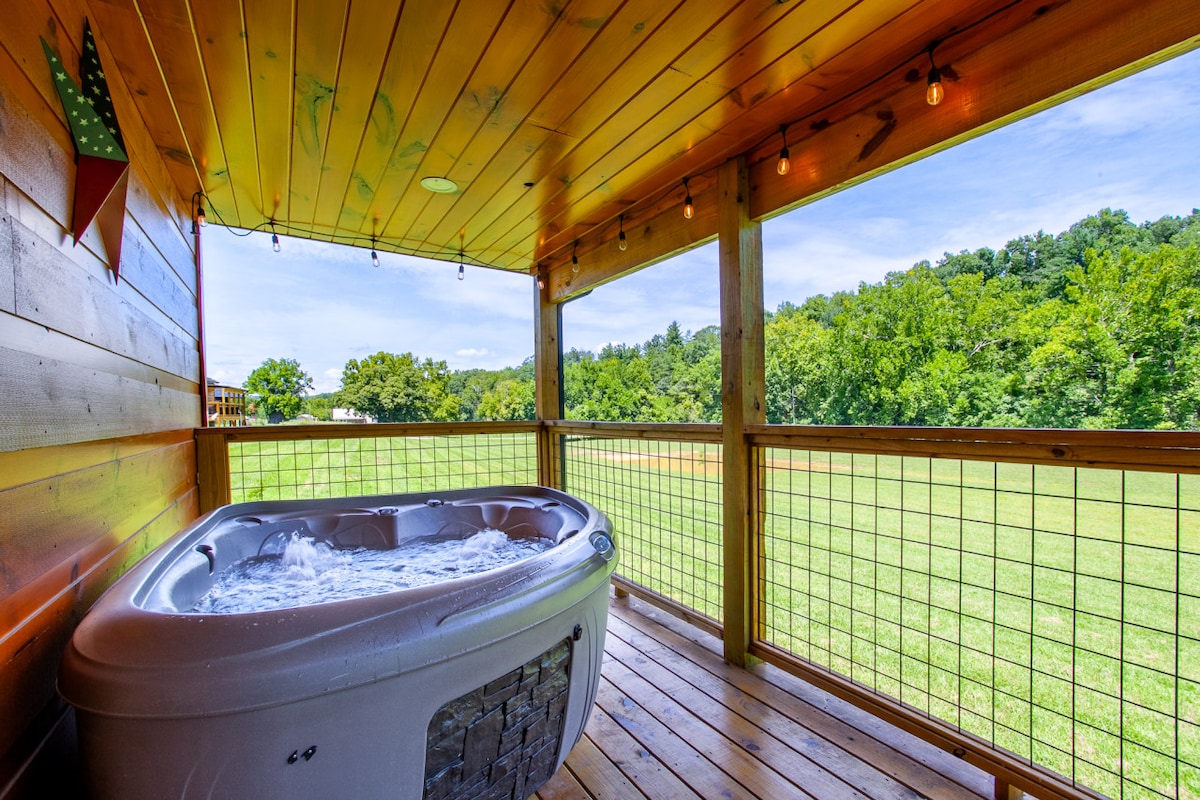 A wooden deck features a hot tub surrounded by rustic wooden railings. Soft string lights enhance the ambiance, while lush green trees and open fields create a serene backdrop under a clear sky.