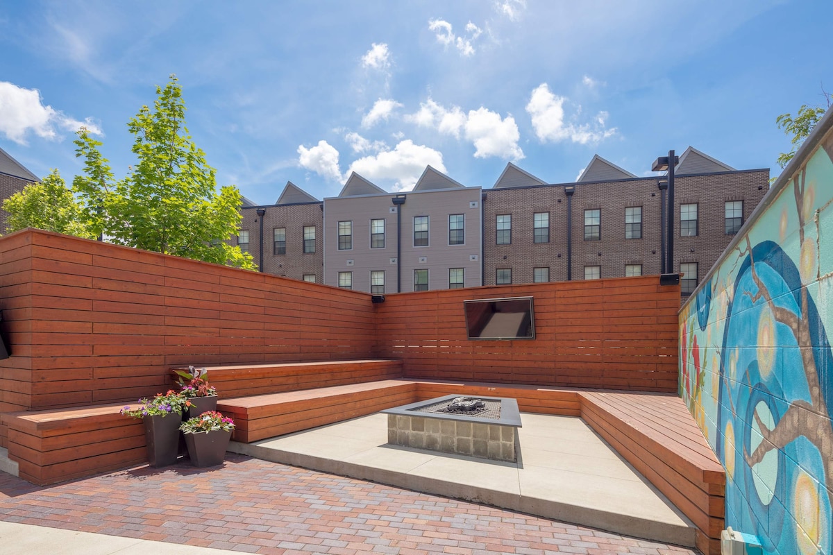 An outdoor gathering space features tiered wooden seating surrounding a stone fire pit, positioned on a paved area. Vibrant greenery is present in planters, and a colorful mural adorns one wall. The backdrop includes a multi-story brick building under a clear blue sky.