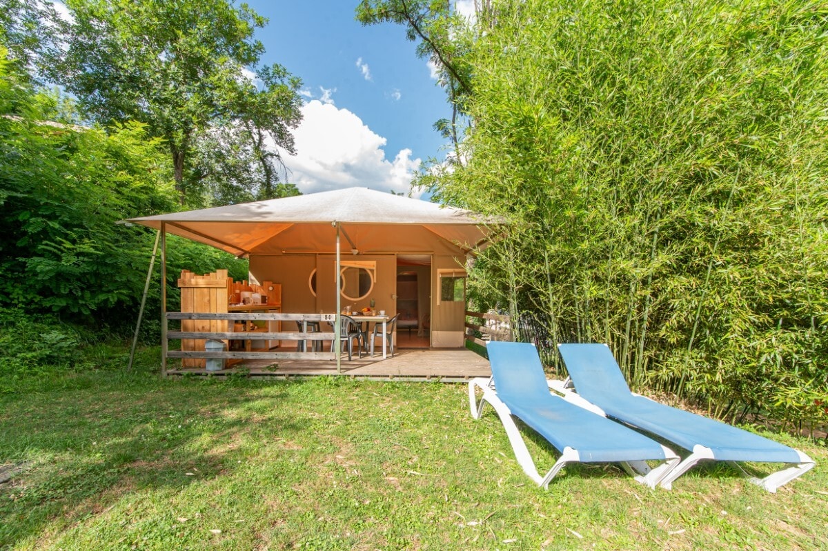 An outdoor seating area is presented with two sun loungers against a backdrop of lush greenery. A covered terrace features a dining space, while a bright sky and fluffy clouds enhance the inviting atmosphere. The entrance to the lodge is visible, creating a cozy retreat.
