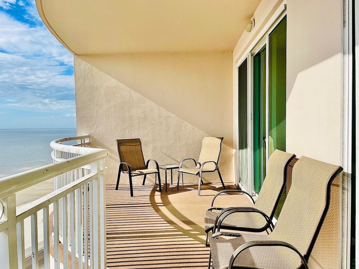 A private balcony on the 11th floor features comfortable lounge chairs and a small table. The expansive ocean views are complemented by sunlight casting soft shadows across the deck. Glass doors lead into the living area, enhancing the connection with the outdoors.