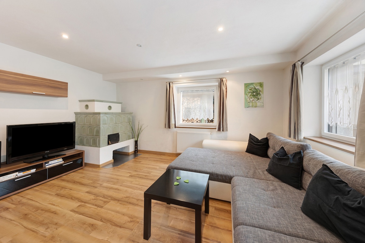 A spacious living room features a comfortable sofa with a chaise lounge, positioned near a window with light curtains. A green tiled fireplace adds a cozy element, while a flat-screen TV is placed on a low shelving unit. Natural wood flooring contributes to a warm ambiance.