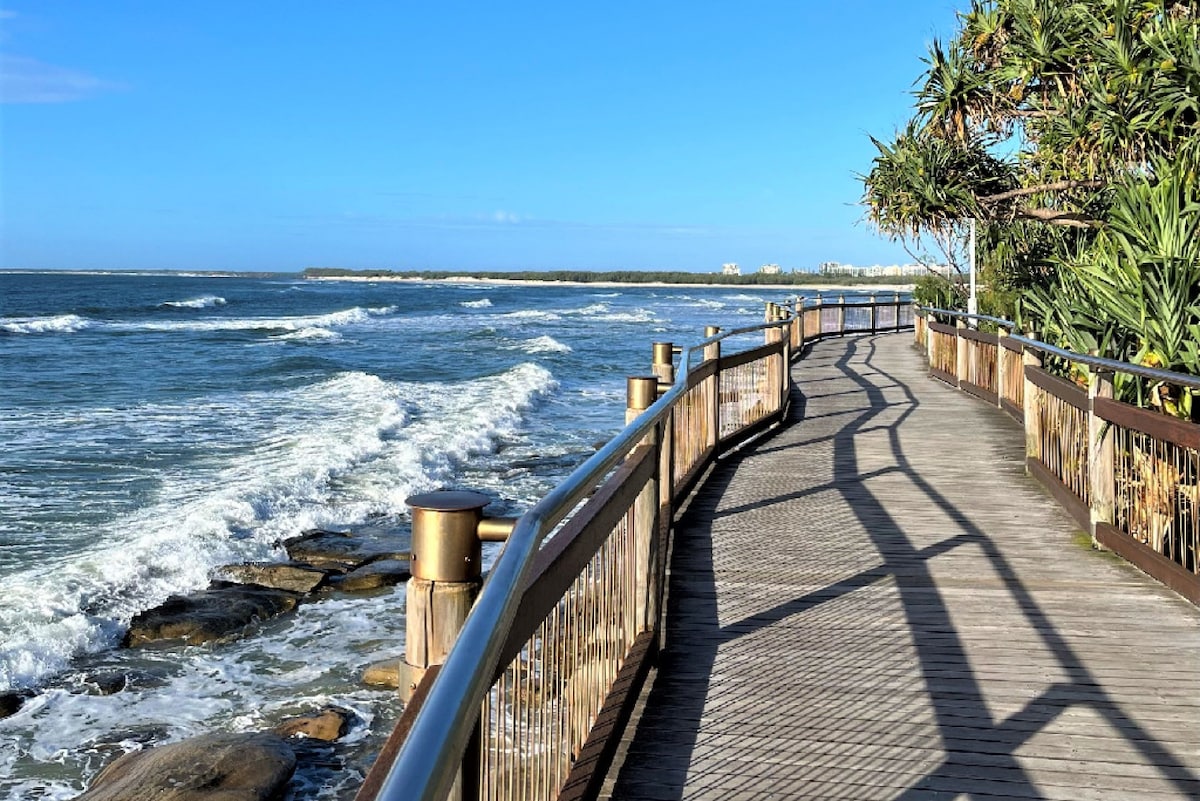 A wooden boardwalk winds alongside the ocean, with gentle waves lapping against the rocky shore. Lush tropical vegetation borders the path, and a clear blue sky stretches overhead, creating a serene coastal scene.