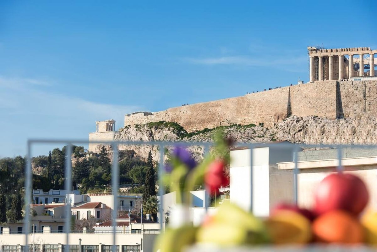 A panoramic view of the Acropolis is presented, showcasing its ancient structures against a clear blue sky. In the foreground, a colorful arrangement of fresh fruits can be seen, providing a vibrant contrast to the historic backdrop.