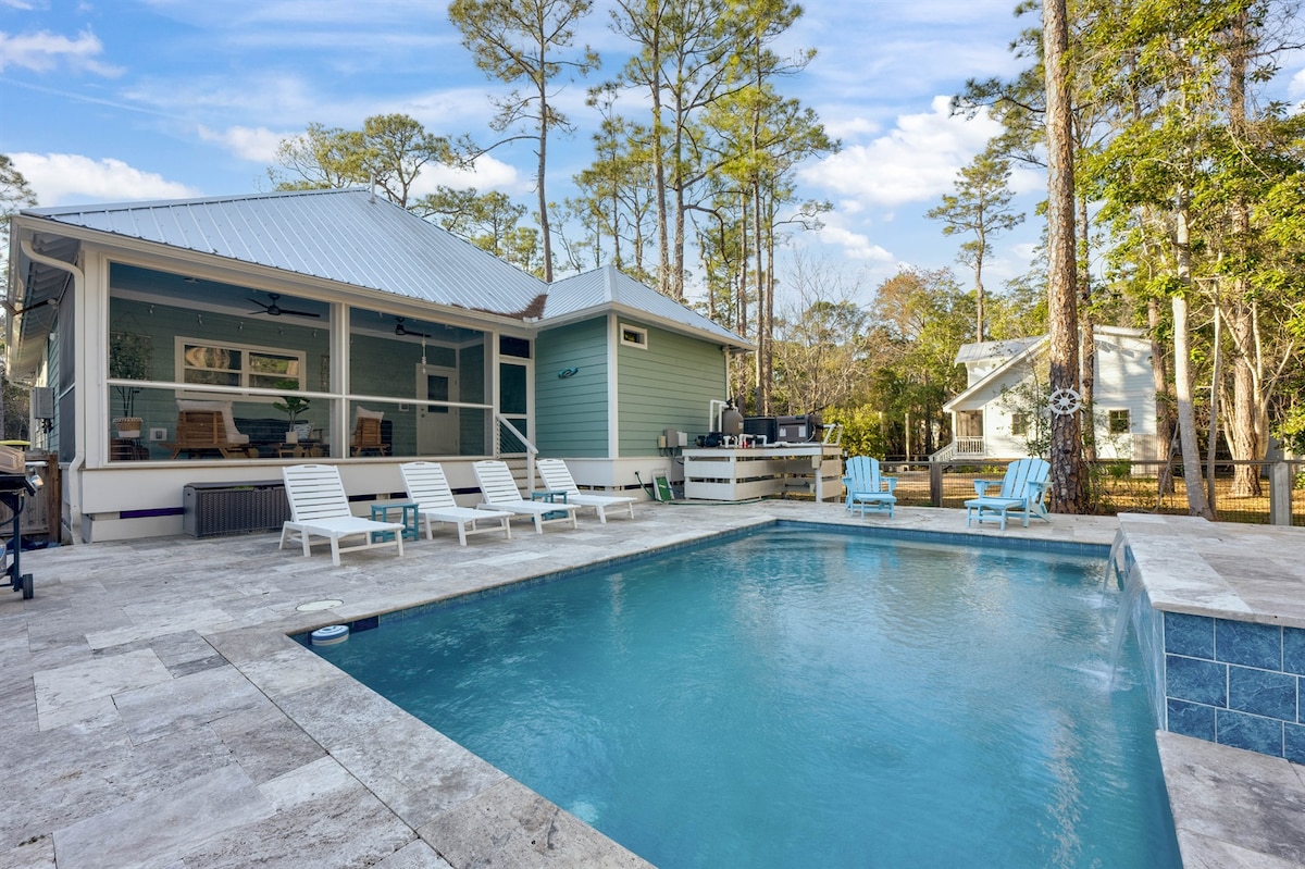 An outdoor pool area features a spacious pool with a waterfall and custom lighting. Surrounding the pool are several lounge chairs positioned for relaxation. On the left, a screened-in porch provides a view of the pool, with trees and a quiet neighborhood visible in the background.