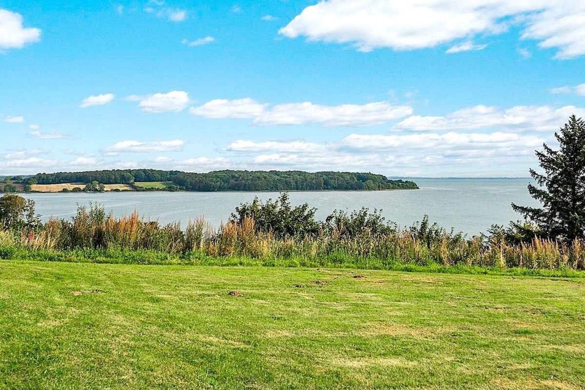 A panoramic view captures a serene stretch of water, bordered by lush greenery and trees. Soft, rolling hills can be seen in the distance, while fluffy clouds drift across a bright blue sky, enhancing the tranquil landscape.