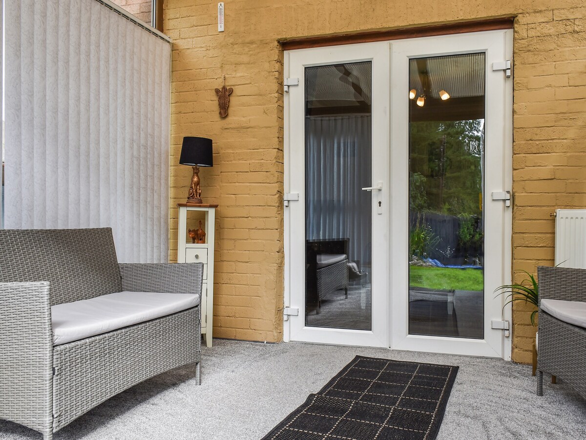 A bright conservatory features two wicker chairs with soft cushions and a small table. Large glass double doors open to the garden, allowing natural light to flow in. A decorative lamp and wall art add warmth to the space, which is finished with a soft carpet.