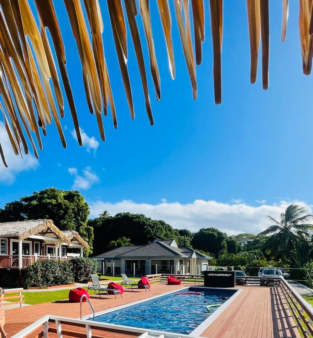 The pool area is framed by a wooden deck and features lounge chairs in a vibrant red hue. A covered outdoor dining area and barbecue grill are positioned nearby. Lush greenery and a clear blue sky create a serene backdrop, enhancing the tropical setting.