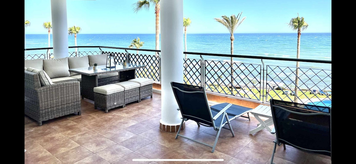 An outdoor terrace features wicker furniture and sun loungers, with a backdrop of the sea and palm trees swaying in the breeze. The view highlights the coastline and clear blue sky, offering a serene space for relaxation.