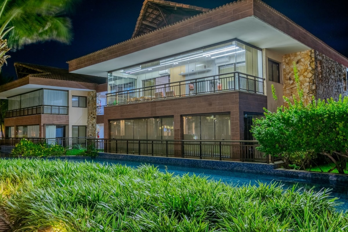 A two-story building is shown at night, illuminated by exterior lights. Expansive glass windows offer a view of the interior, while lush greenery borders the property's edge. A tranquil pool reflects the surrounding plants and architecture, enhancing the inviting ambiance of the area.