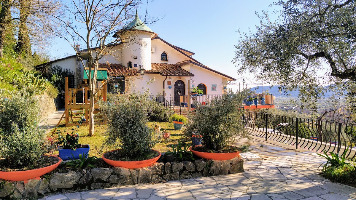 The exterior of Villa Vernazza is surrounded by lush greenery and a well-maintained garden. The charming structure features a round turret and a terracotta-tiled roof. Plant pots filled with vibrant flowers line the pathway, leading to a welcoming entrance illuminated by natural light.