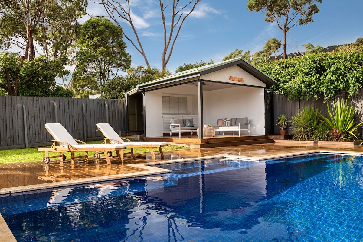 A spacious outdoor area features a crystal-clear pool surrounded by a wooden deck. Two lounge chairs invite relaxation, while a poolside cabana offers additional seating. Lush greenery lines the perimeter, providing a serene backdrop for outdoor enjoyment.
