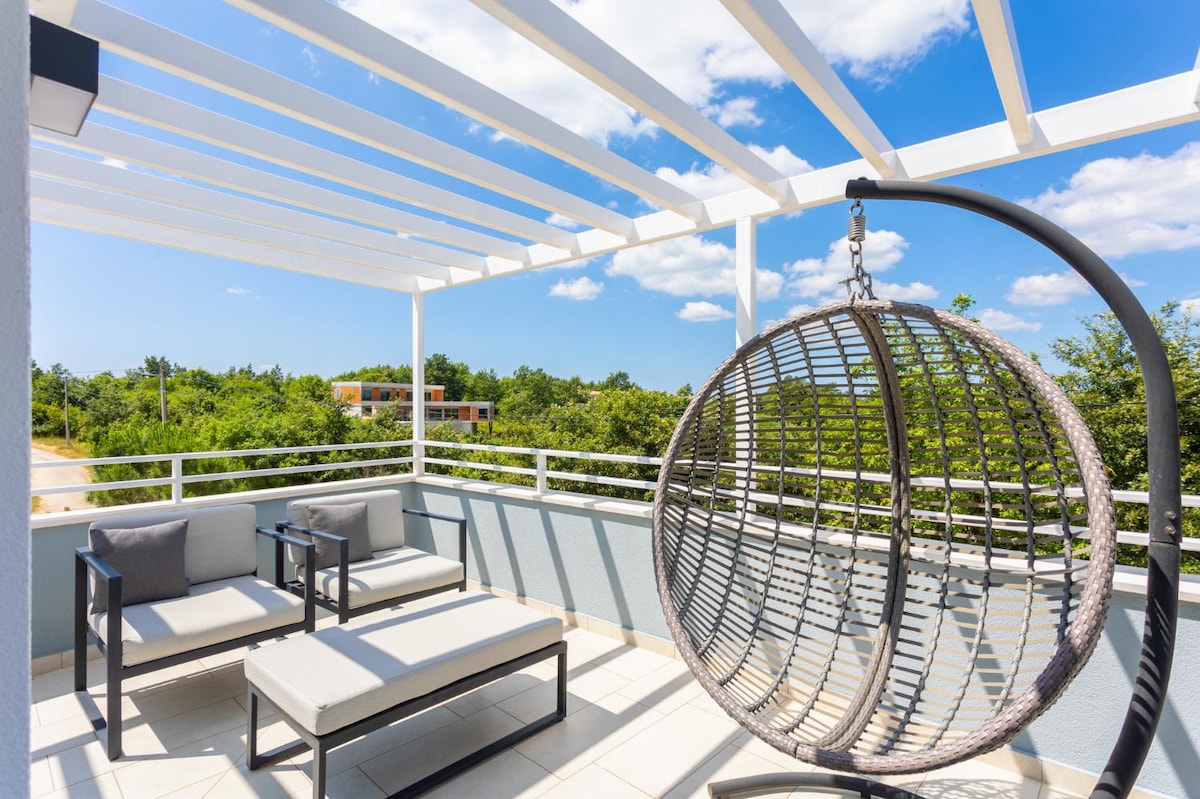 A spacious terrace features a hanging chair and comfortable seating arrangement, offering views of lush greenery. A sunlit atmosphere is provided by the open space and modern design, with light-colored furniture complementing the surrounding landscape.